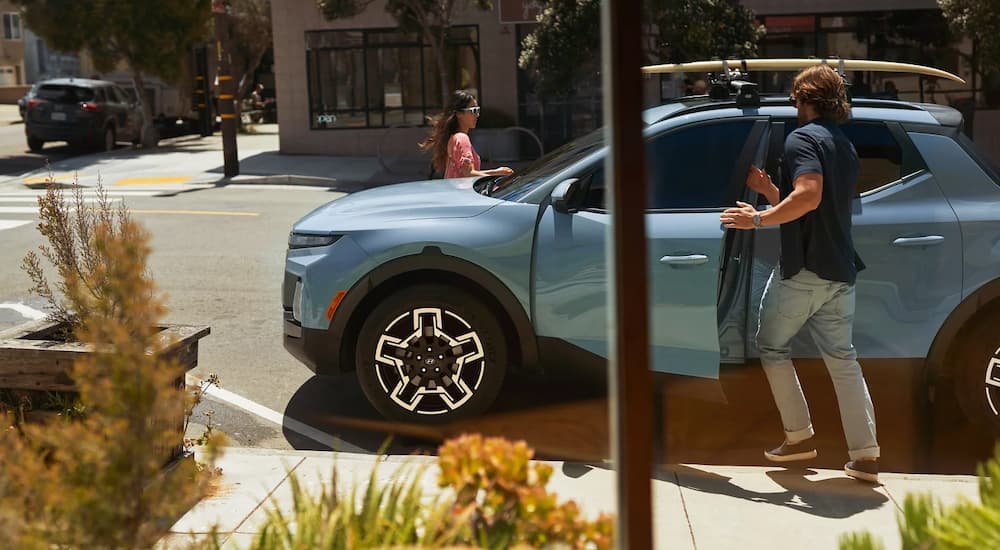A man is getting into a blue 2025 Hyundai Santa Cruz with a surfboard on the roof.