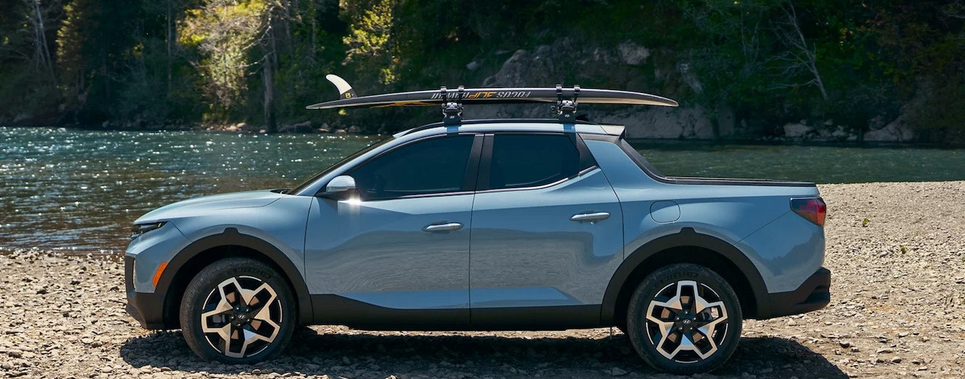 A blue 2024 Hyundai Santa Cruz parked near water with a surfboard on the roof.