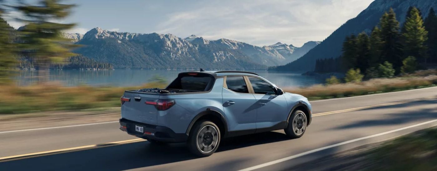 Blue 2026 Hyundai Santa Cruz driving on the road near mountains and a lake.