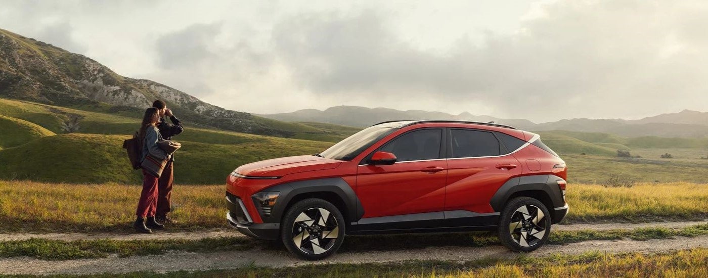 Two people standing in front of a red 2024 Hyundai Kona parked on a trail in a field.