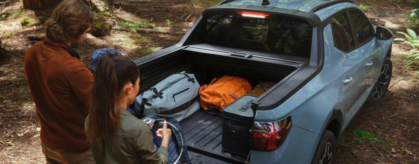 A couple loading camping gear into the bed of a blue 2025 Hyundai Santa Cruz Limited in the woods.